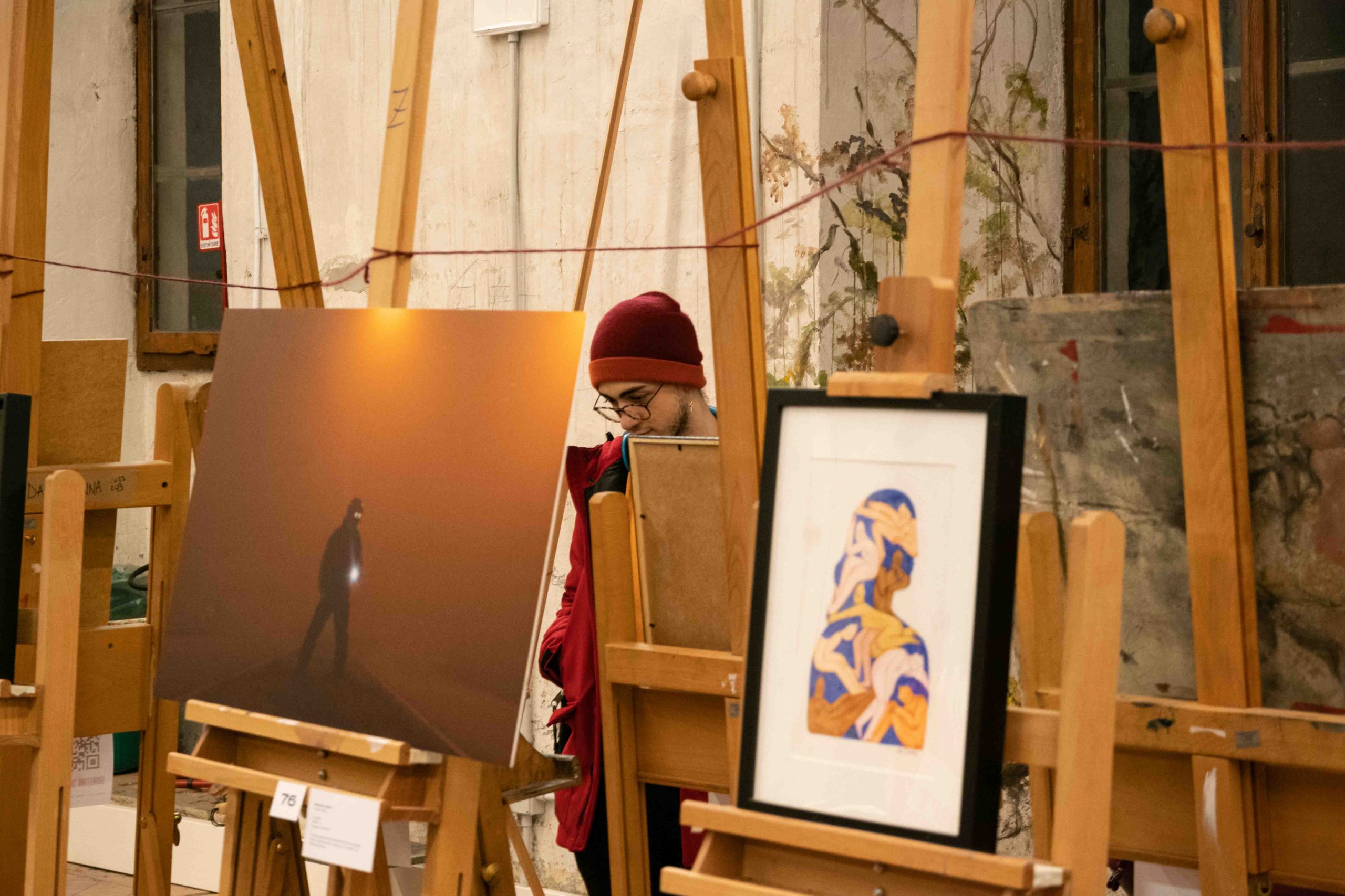 Quadri su cavalletti in una galleria artistica con una persona sullo sfondo che osserva le opere esposte.