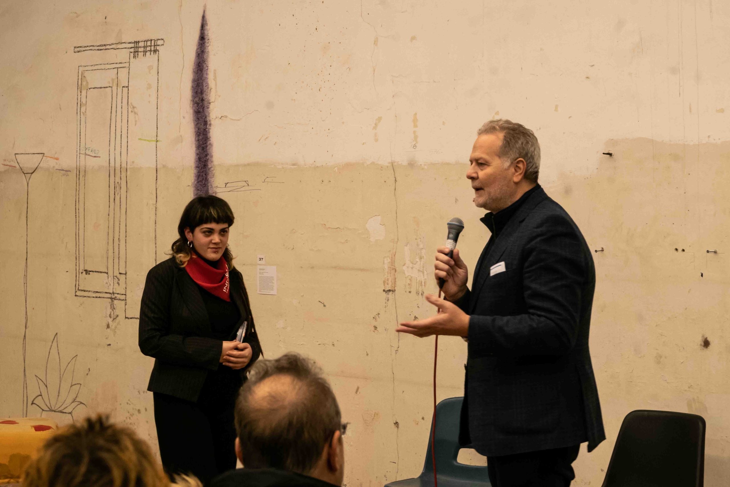 Due persone parlano davanti a un pubblico in una sala con murales. Una donna ascolta mentre un uomo parla al microfono.