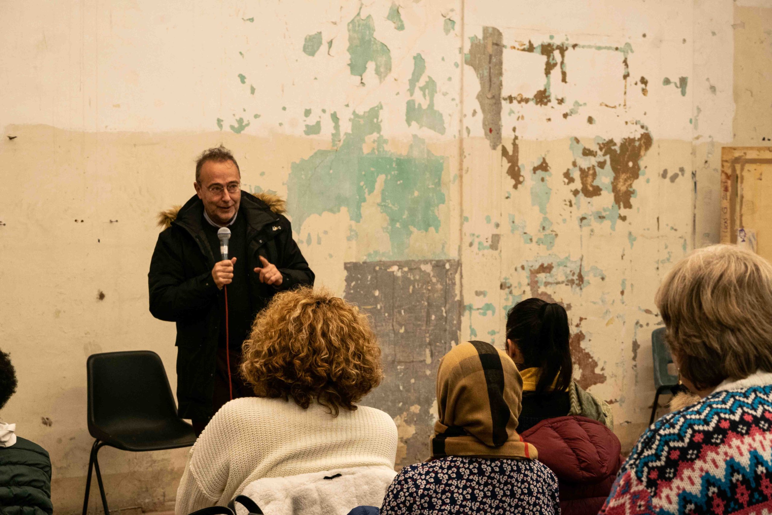 Persona parla al pubblico in una sala, con pareti usurate, coinvolgendo figure sedute che ascoltano attentamente.