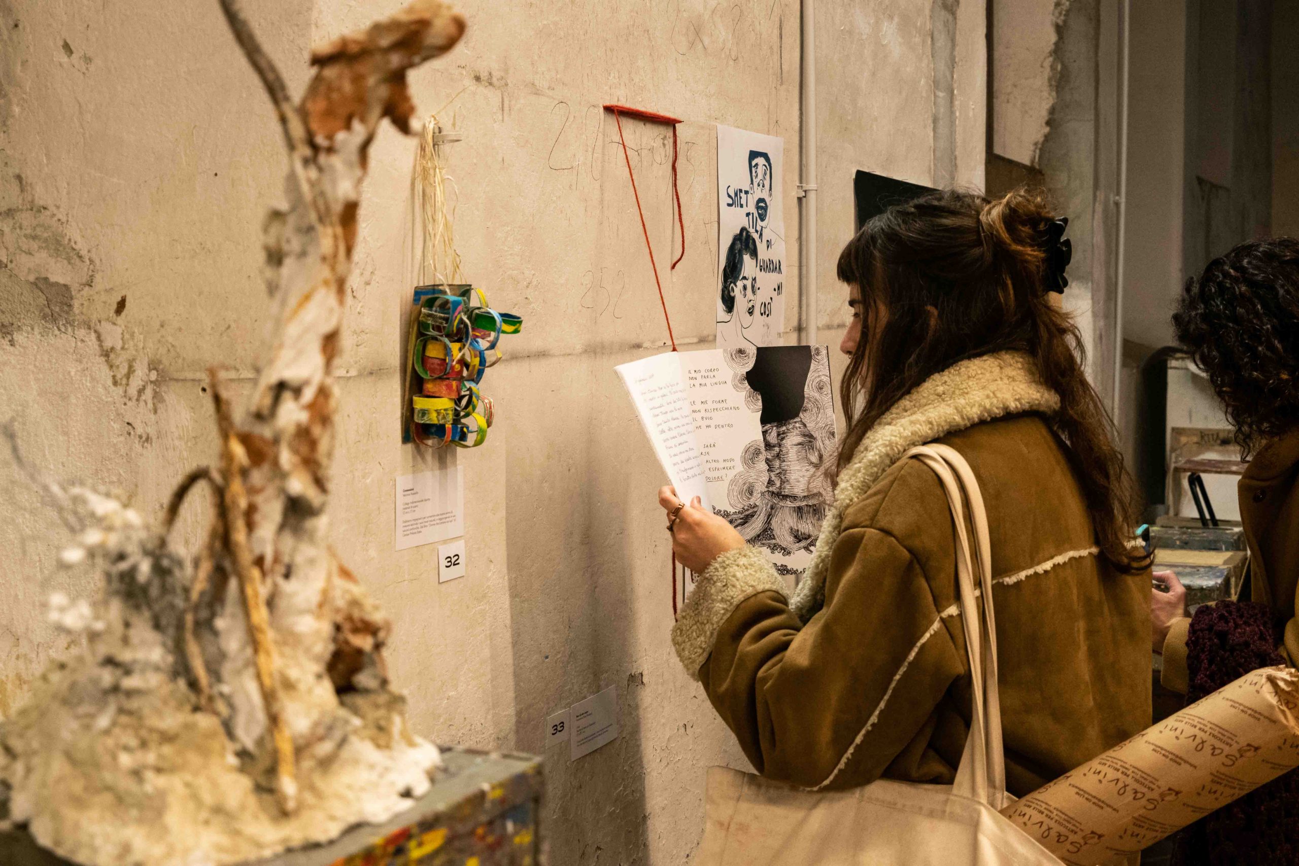 Donna osserva installazione artistica in galleria, leggendo brochure. Opera in primo piano e parete decorata.