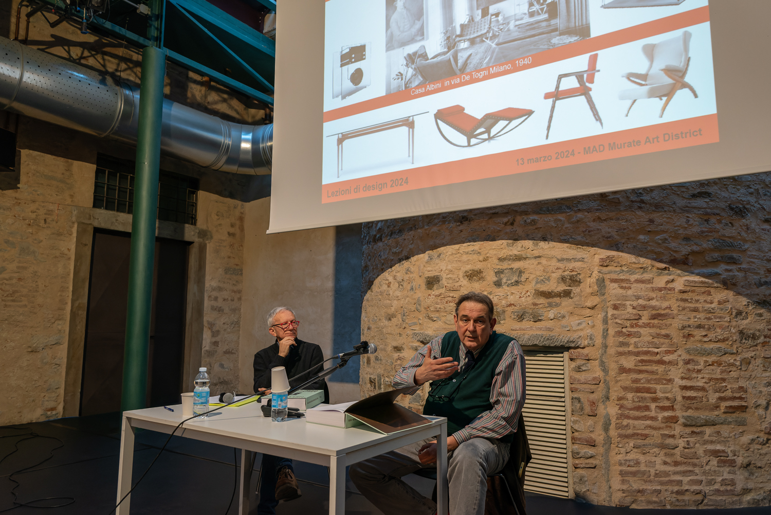 Due uomini discutono di design in una conferenza, con proiezione di immagini di mobili su uno schermo.