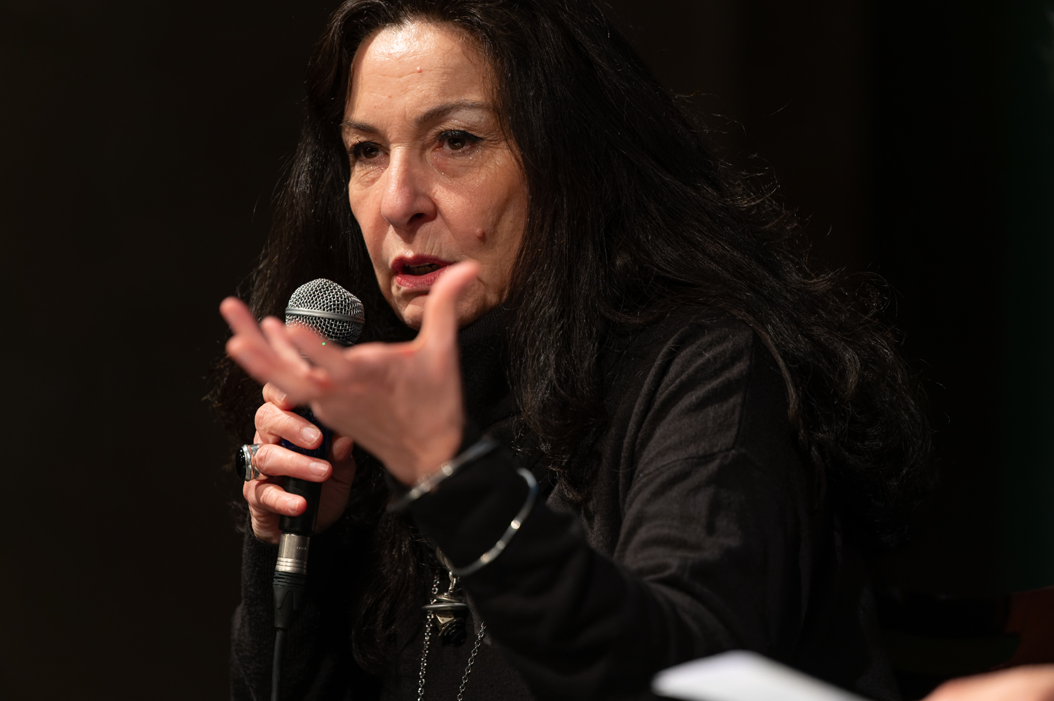 Donna con microfono, gesticola mentre parla, indossa abiti scuri e bracciali, sfondo scuro.