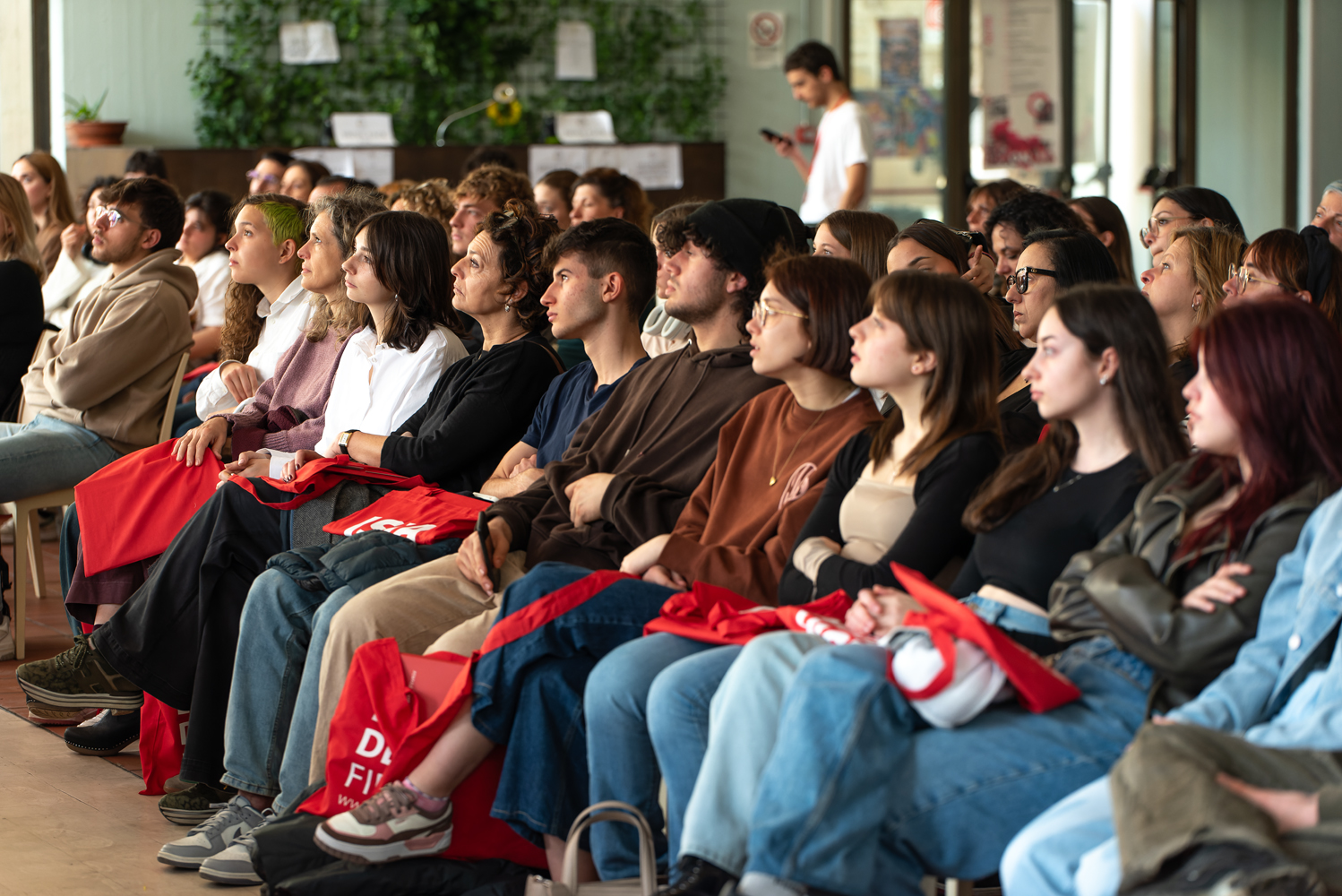 Persone sedute attentamente in un auditorium, con borse rosse, durante un evento o una conferenza.