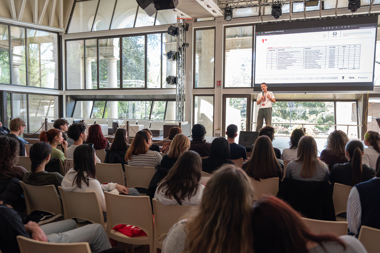 Presentazione a una conferenza con pubblico attento, in una sala con ampie finestre e schermo proiettato.