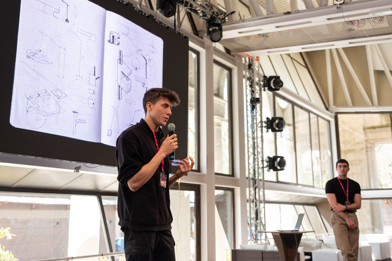 Oratore con microfono illustra progetti su schermo in una conferenza, ambiente moderno con luci e finestre ampie.