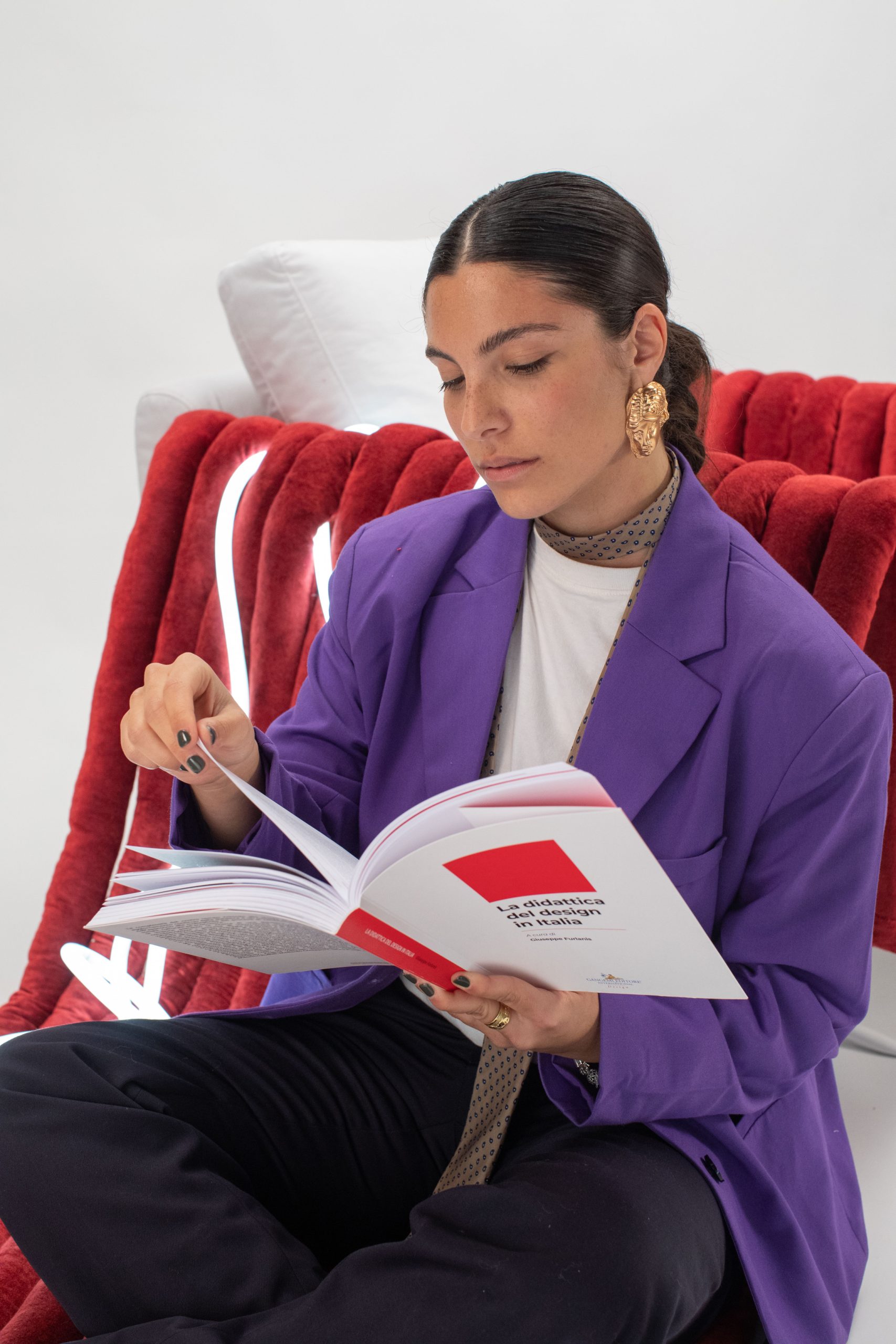 Donna legge un libro su design italiano, seduta su una sedia rossa, vestita in completo viola e gioielli dorati.