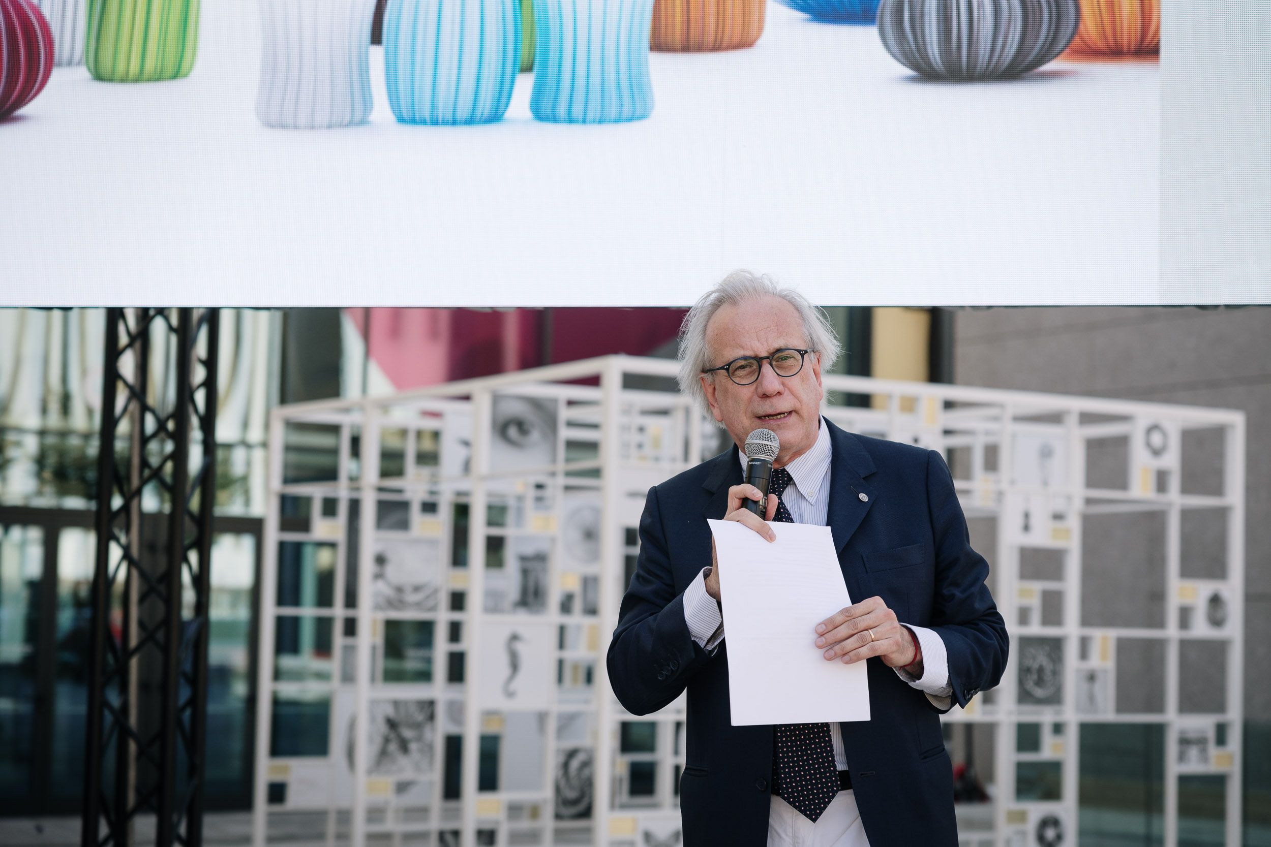 Persona anziana parla al microfono durante un evento all'aperto, tenendo un foglio. Scultura colorata sullo sfondo.