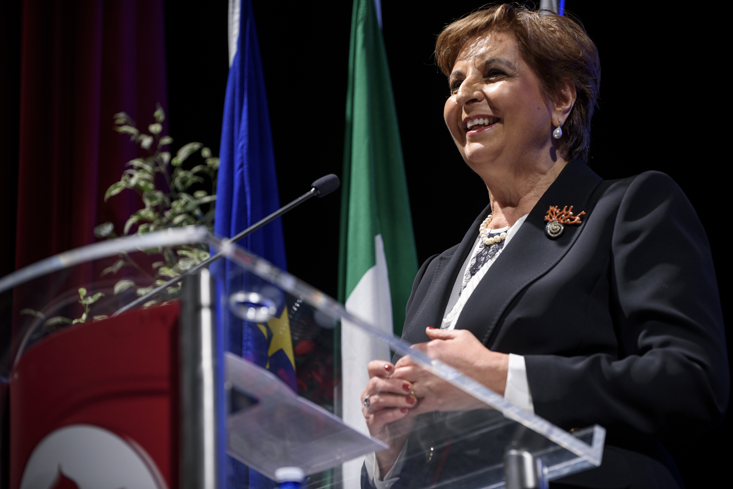 Donna sorridente in abito elegante parla a un podio, sfondo con bandiere italiane ed europee, pianta a lato.