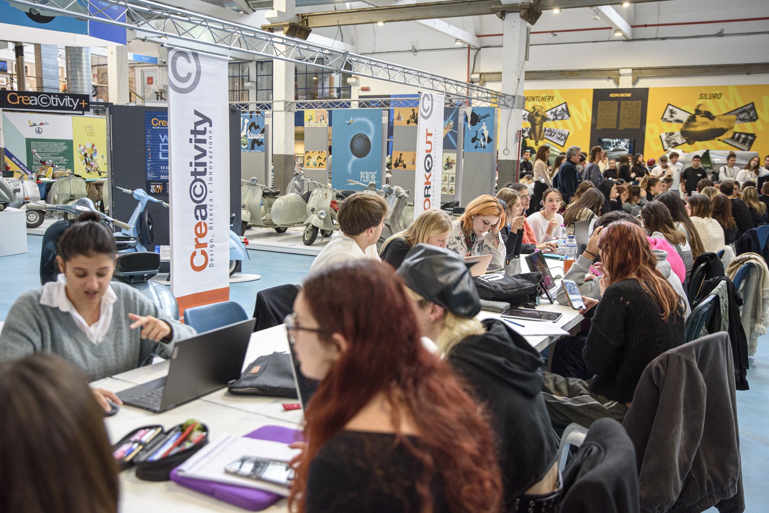 Persone lavorano ai computer in un evento di creatività, circondati da poster e Vespe esposte.