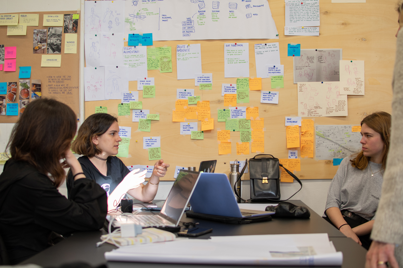 Tre persone discutono di fronte a una lavagna piena di note adesive colorate in un ufficio creativo.