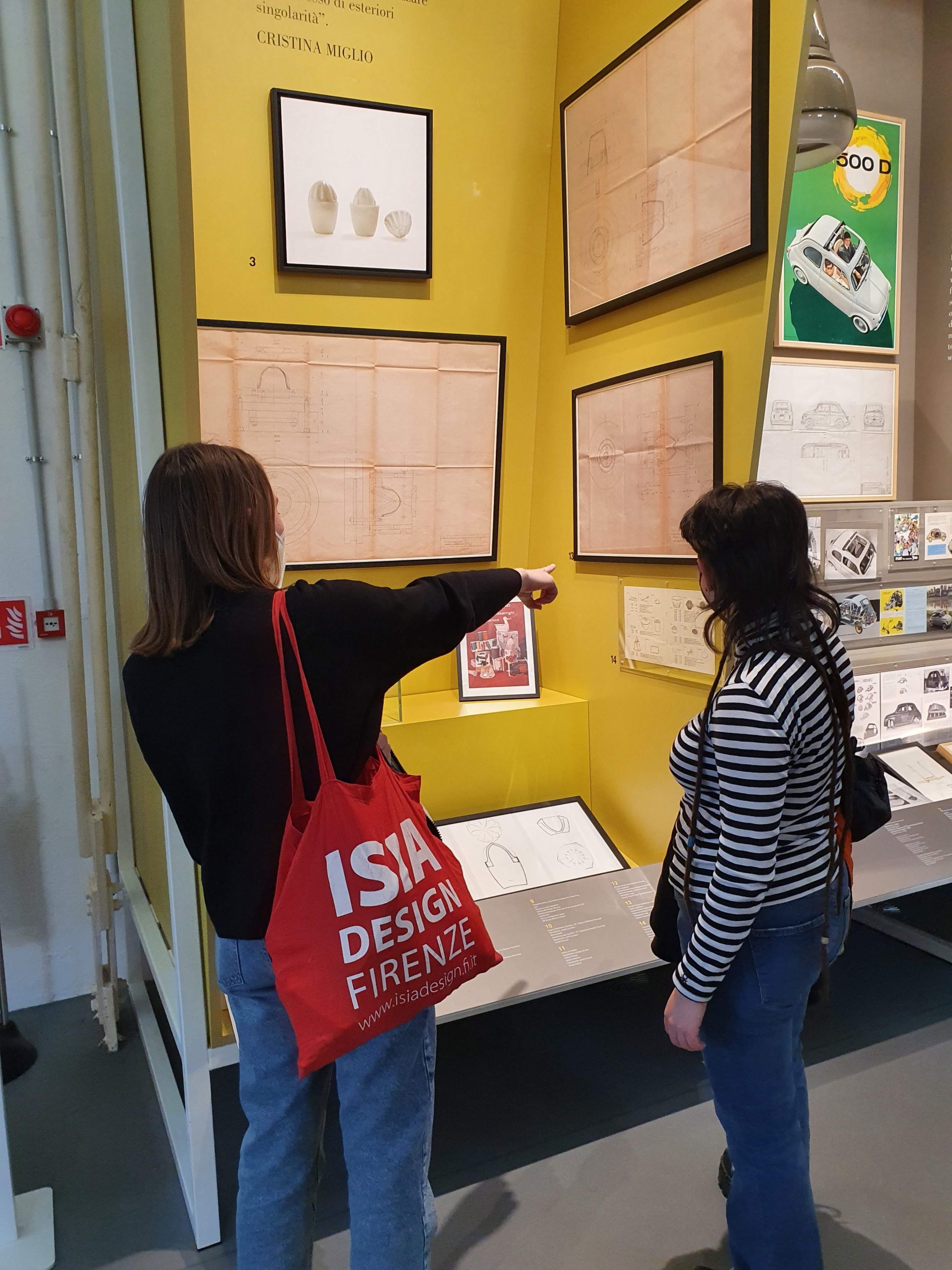 Due persone osservano disegni tecnici in una mostra di design. Uno di loro indica un dettaglio.