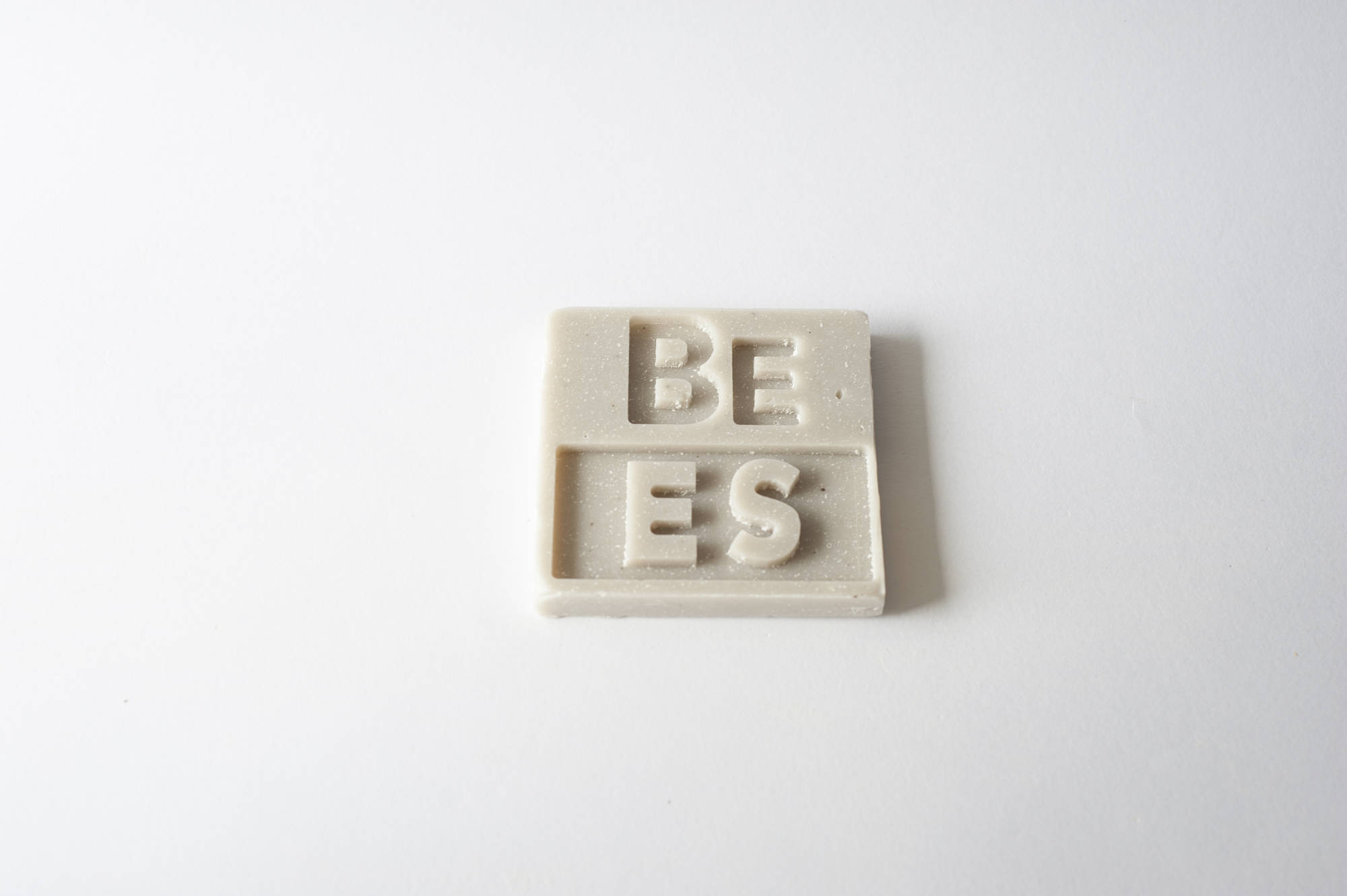 Tessera quadrata con lettere in rilievo BE e ES su sfondo bianco minimalista.