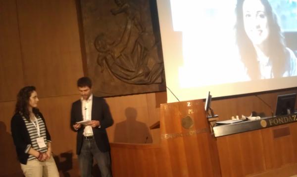 Due persone parlano su un palco durante una presentazione, con un grande schermo dietro e una scultura sul muro.