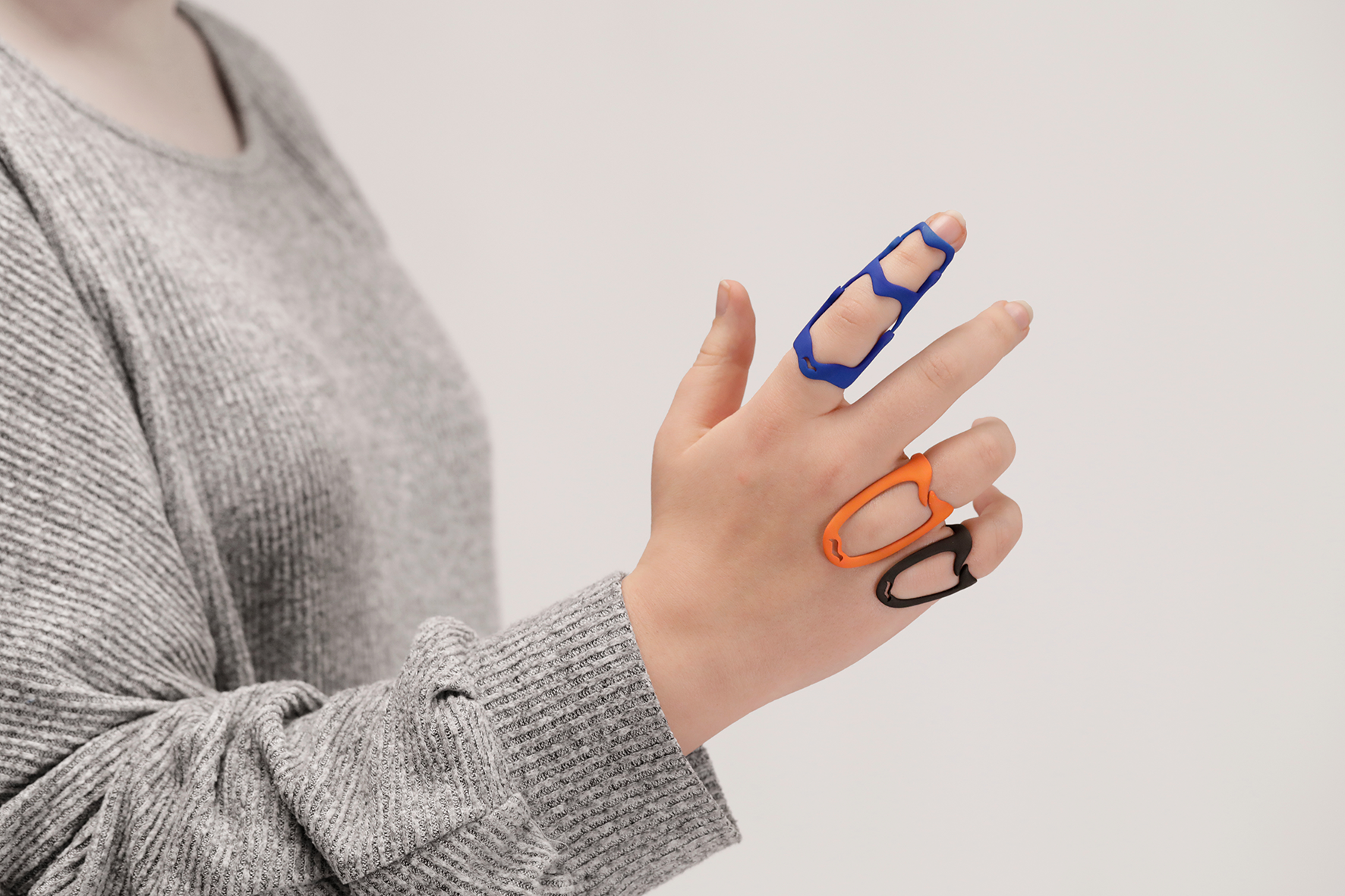 Mano con anelli in plastica colorati su dita, indossa un maglione grigio, sfondo bianco.