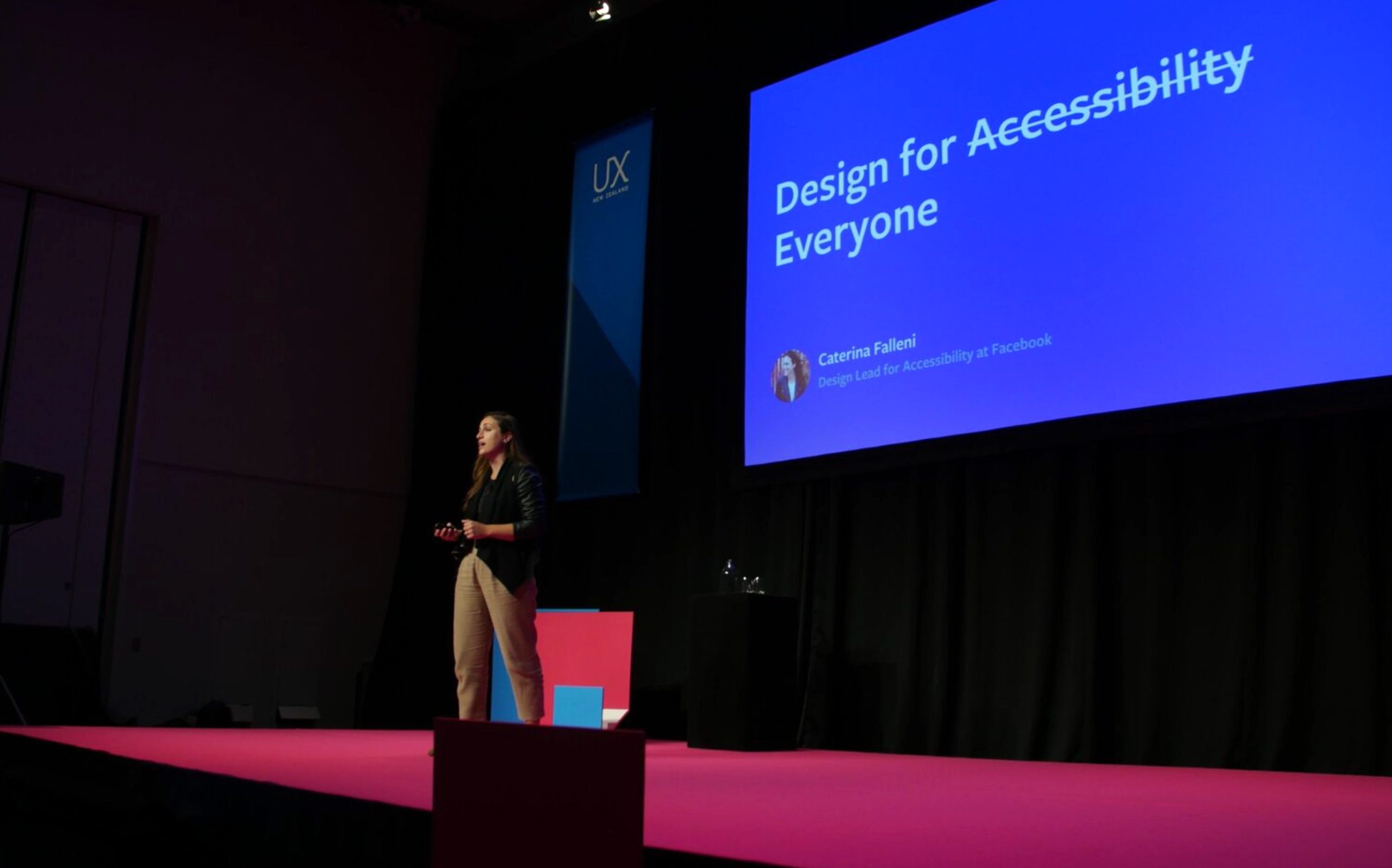 Relatrice su palco parla di design per l'accessibilità a una conferenza UX, schermo blu con testo di apertura.