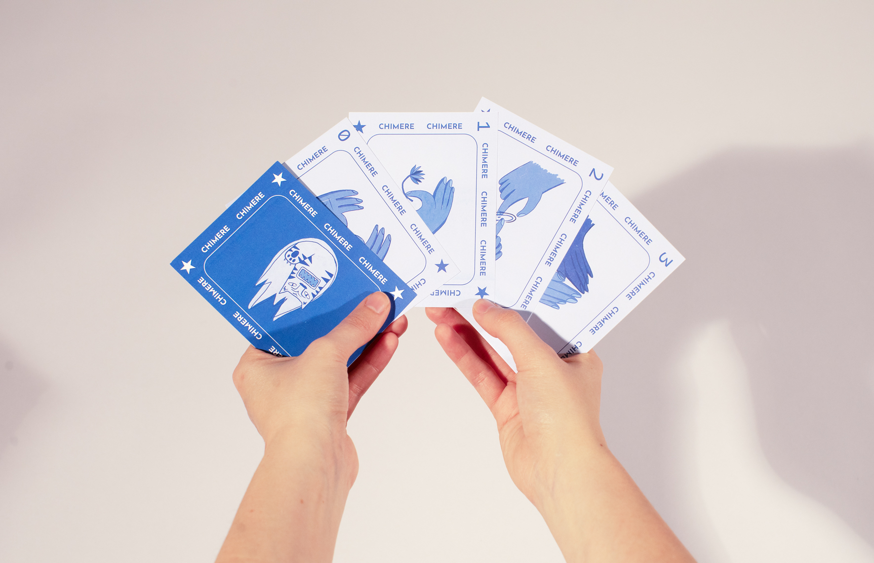 Mani che tengono carte da gioco blu e bianche con scritte Chimere e illustrazioni di mani e figure astratte.