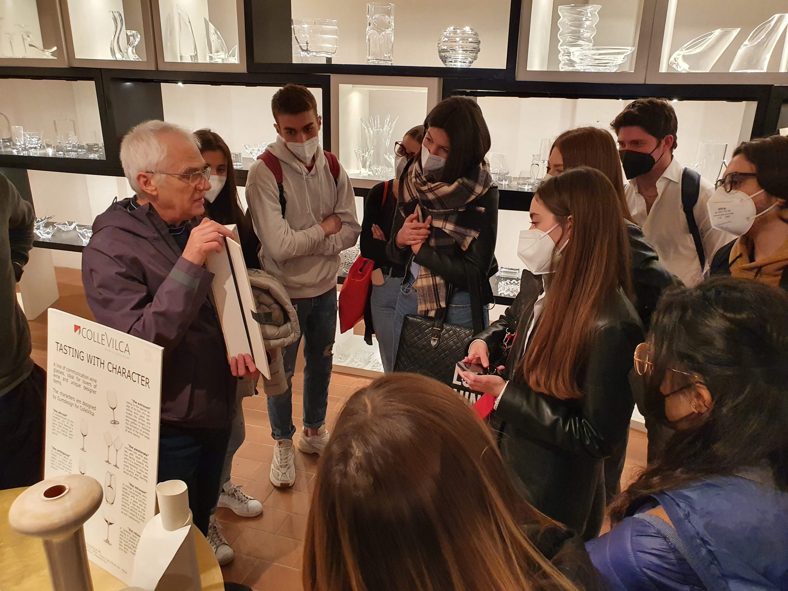 Un gruppo di persone ascolta un esperto che parla davanti a scaffali di cristalleria, in un ambiente espositivo.