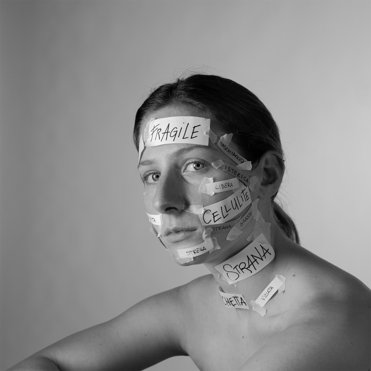 Ritratto in bianco e nero di una donna con parole negative scritte su pezzi di nastro adesivo attaccati al volto.