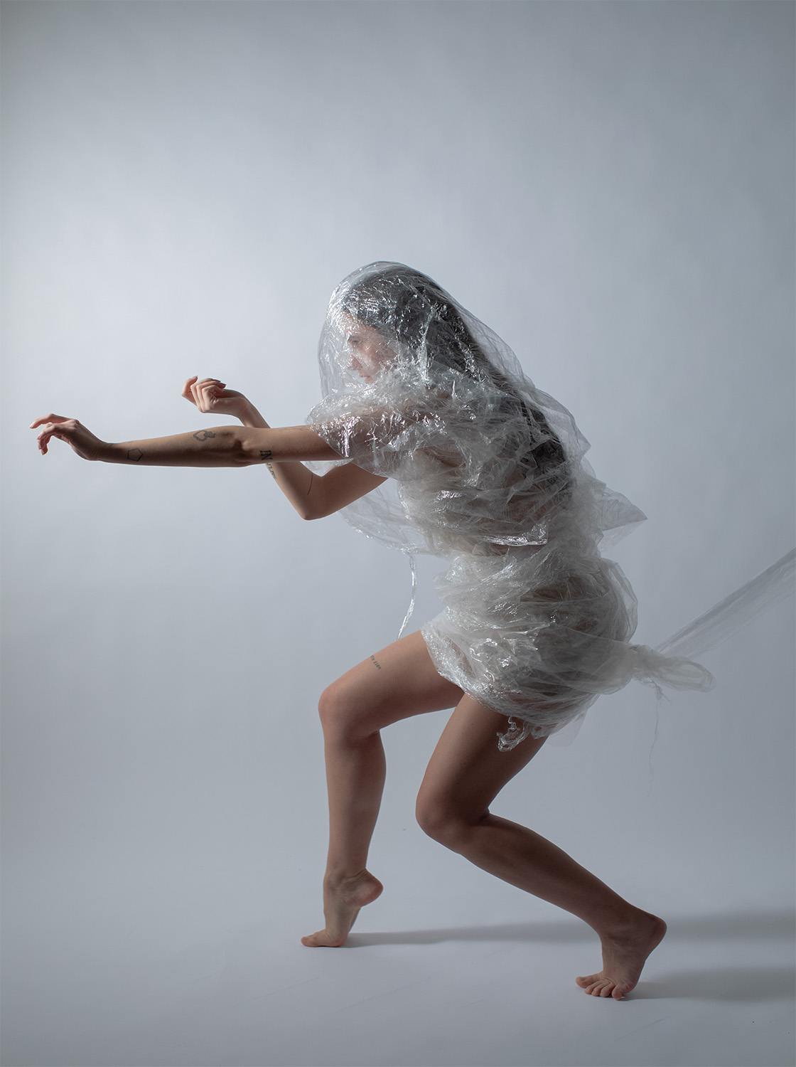 Persona avvolta in plastica trasparente in posa su sfondo neutro, arte concettuale e movimento.