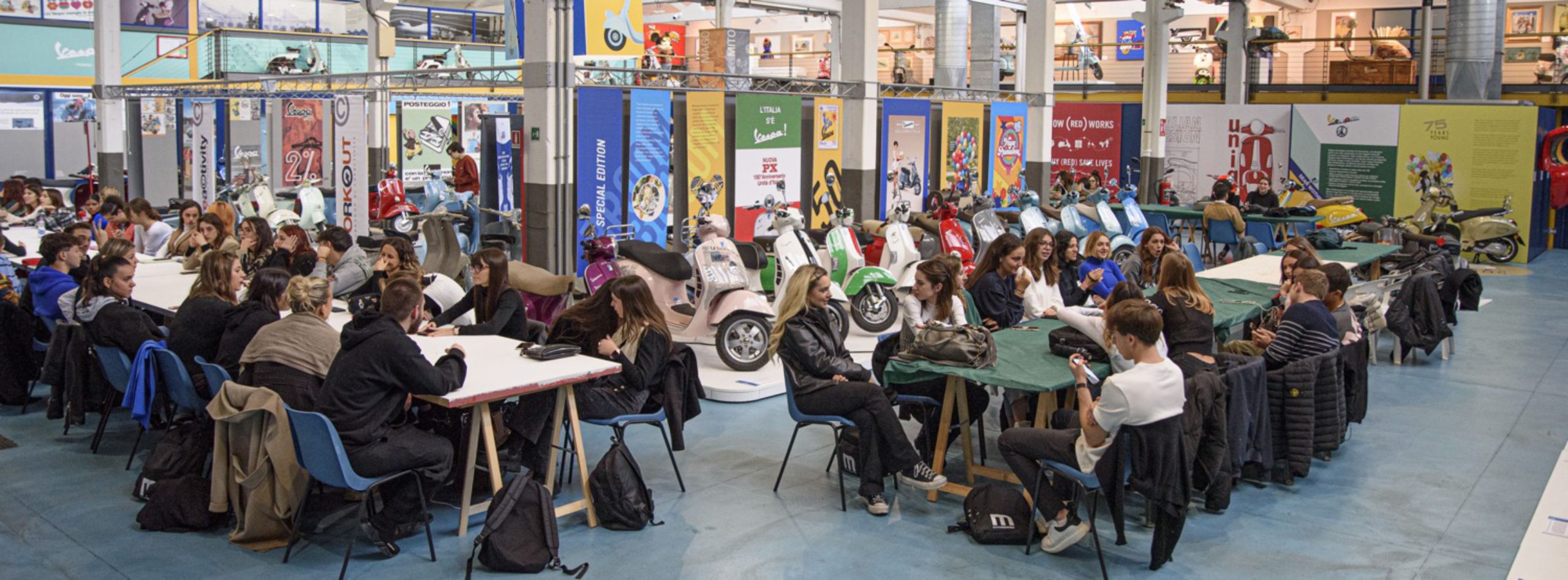 Giovani partecipano a un workshop in un museo, circondati da Vespe e cartelloni colorati.