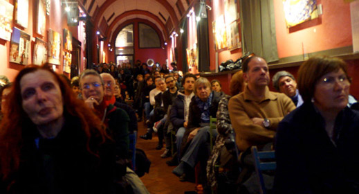 Pubblico attento in una galleria d'arte con pareti rosse, seduto a seguire una presentazione o evento culturale.