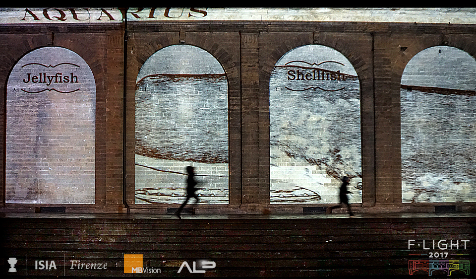 Proiezione artistica su parete con scritto Jellyfish e Shellfish, persone in silhouette camminano davanti.