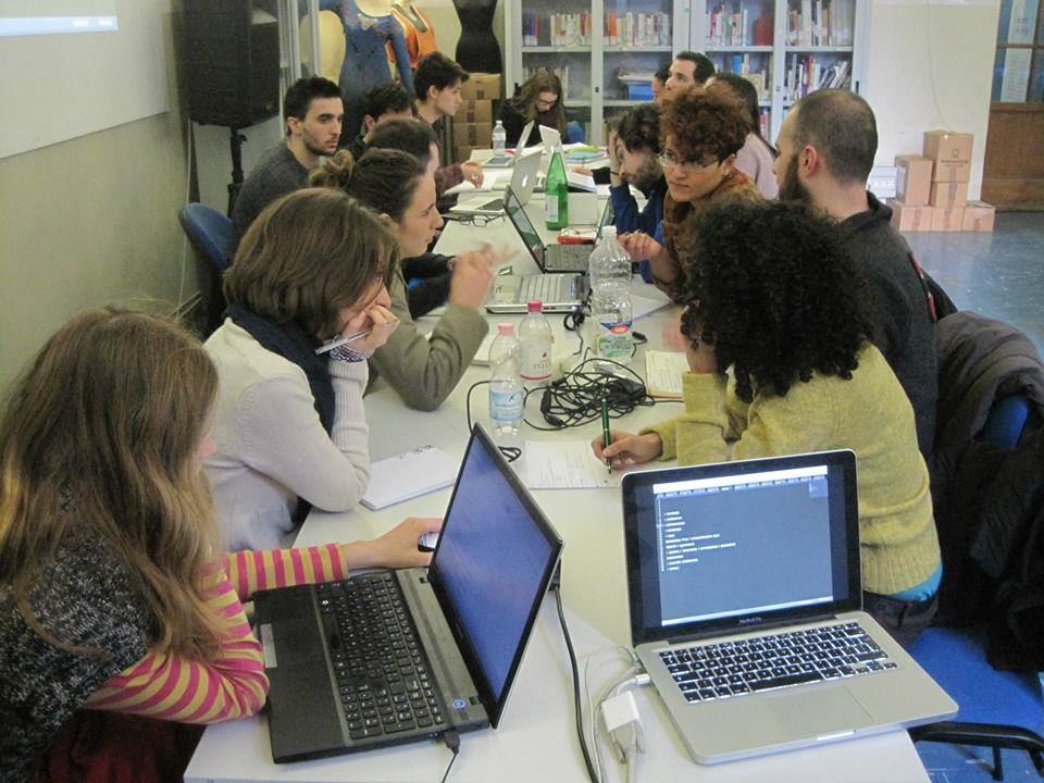 Gruppo di persone attorno a un tavolo collaborano su laptop in una sala con libreria sullo sfondo.