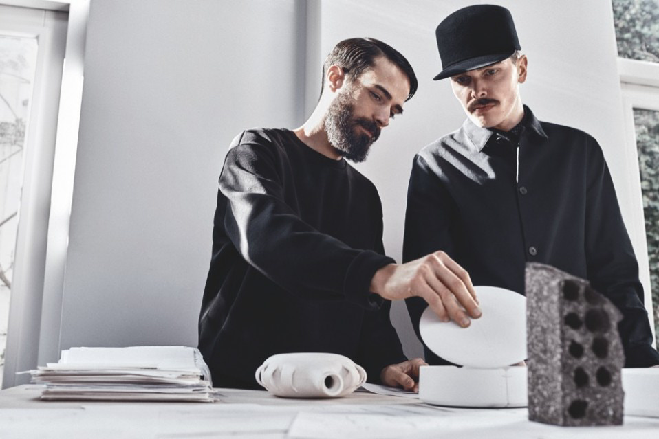 Due uomini discutono un progetto architettonico con modelli di design su un tavolo illuminato dalla luce.