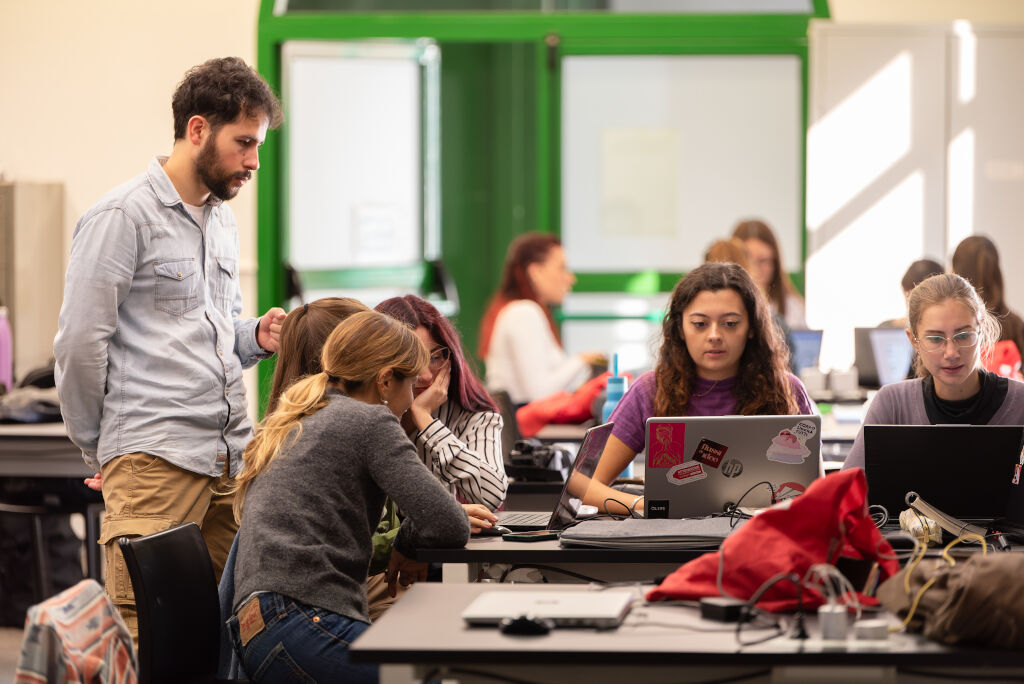 Persone che collaborano in un'aula, lavorando su laptop e discutendo progetti, con un insegnante che guida.