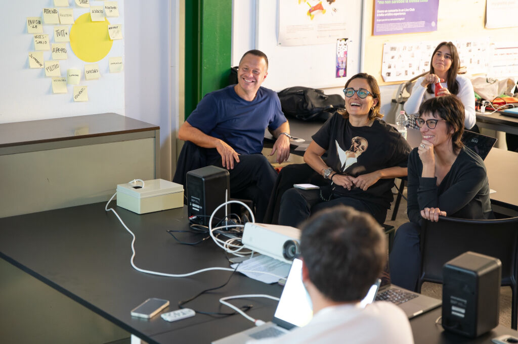 Gruppo di persone sorridenti in riunione, con post-it sul muro e computer in un'aula moderna.