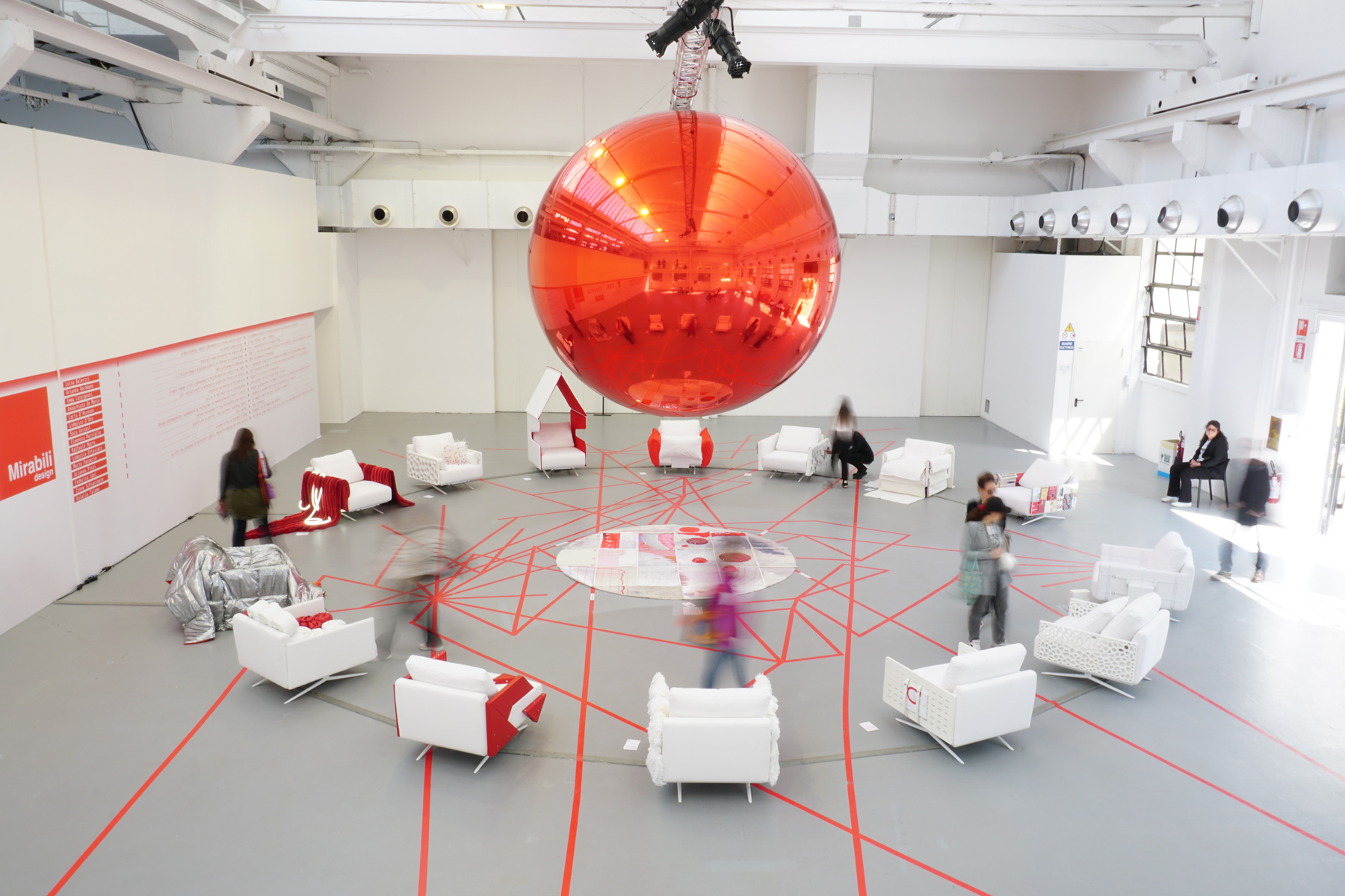 Installazione artistica con grande sfera rossa sospesa in una sala con poltrone bianche e linee rosse sul pavimento.