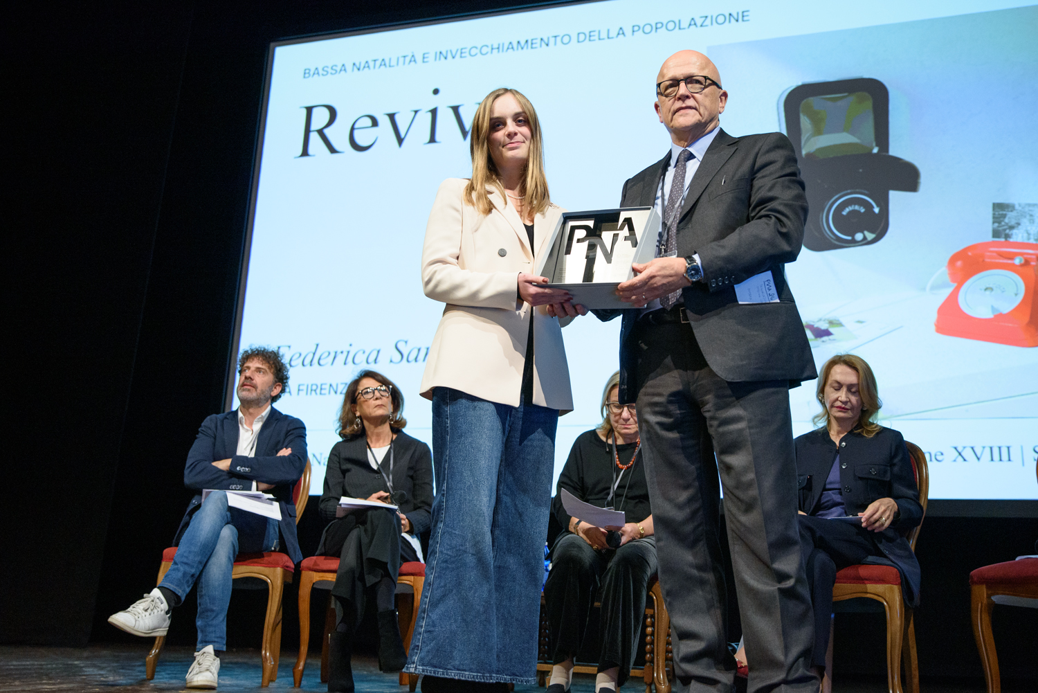 Persone sul palco durante la premiazione di un evento su natalità e invecchiamento, con un trofeo in mostra.