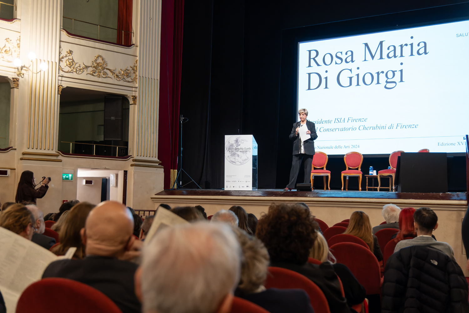 Persona parla sul palco a una conferenza in un auditorium affollato, schermo dietro con testo.