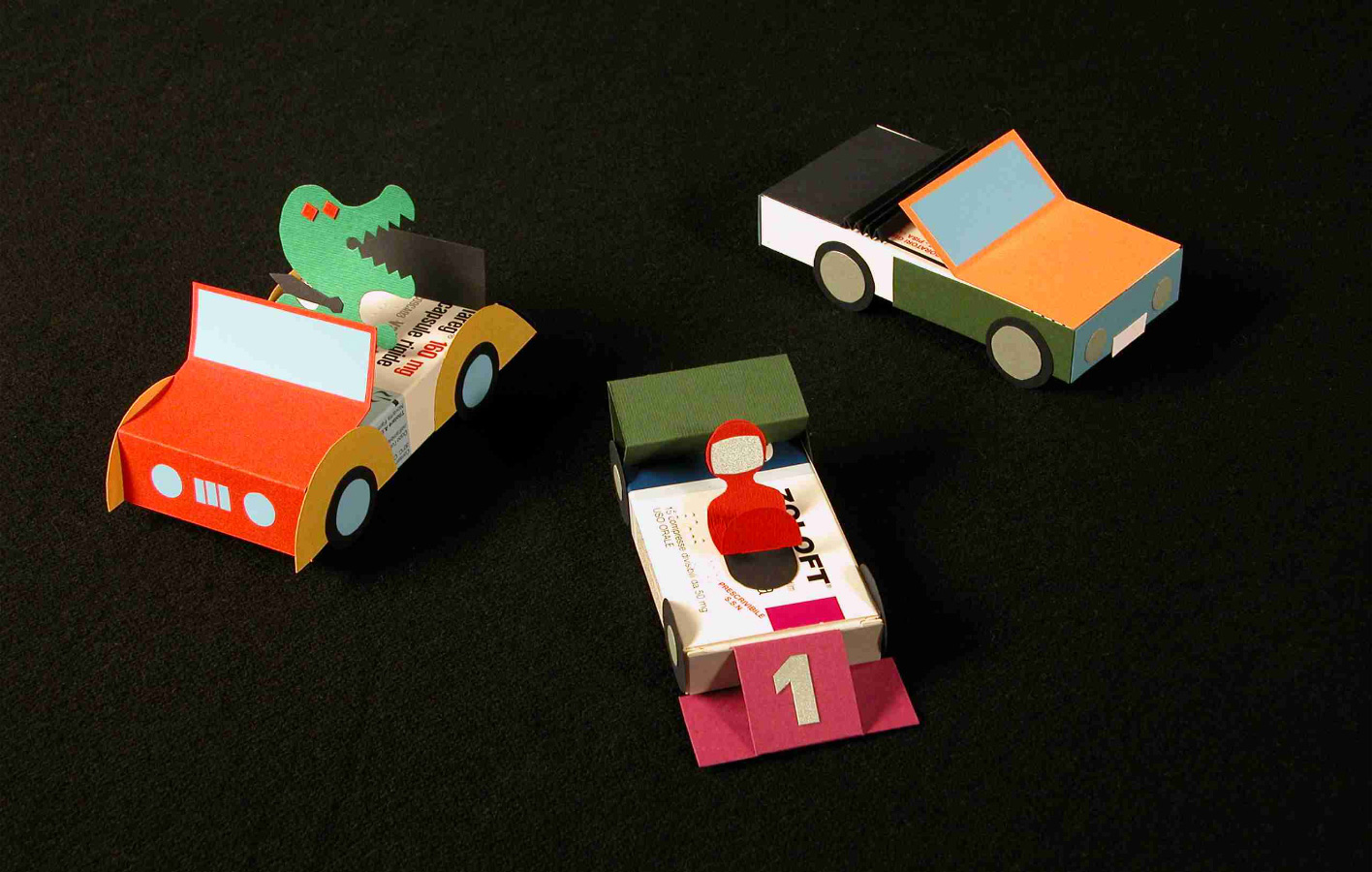 Tre macchinine di carta colorate su sfondo nero, una con un dinosauro, una con un premio numero uno.