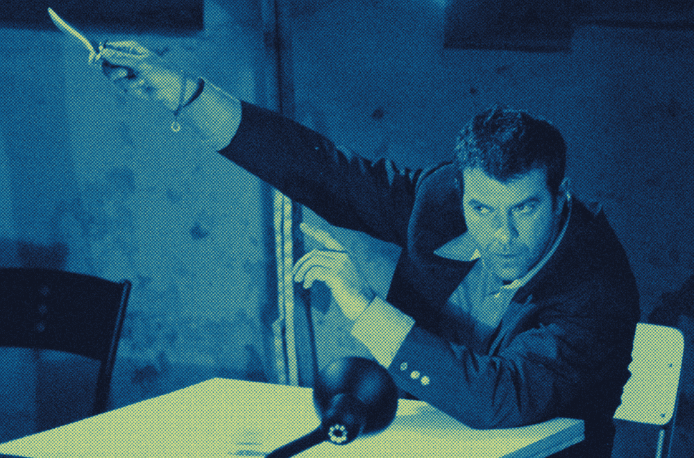 Uomo in giacca scura brandisce un coltello puntato verso l'alto in una stanza illuminata di blu, seduto a un tavolo.