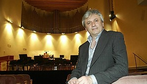 Uomo in abito elegante seduto in un teatro vuoto con arredi e organo sullo sfondo.