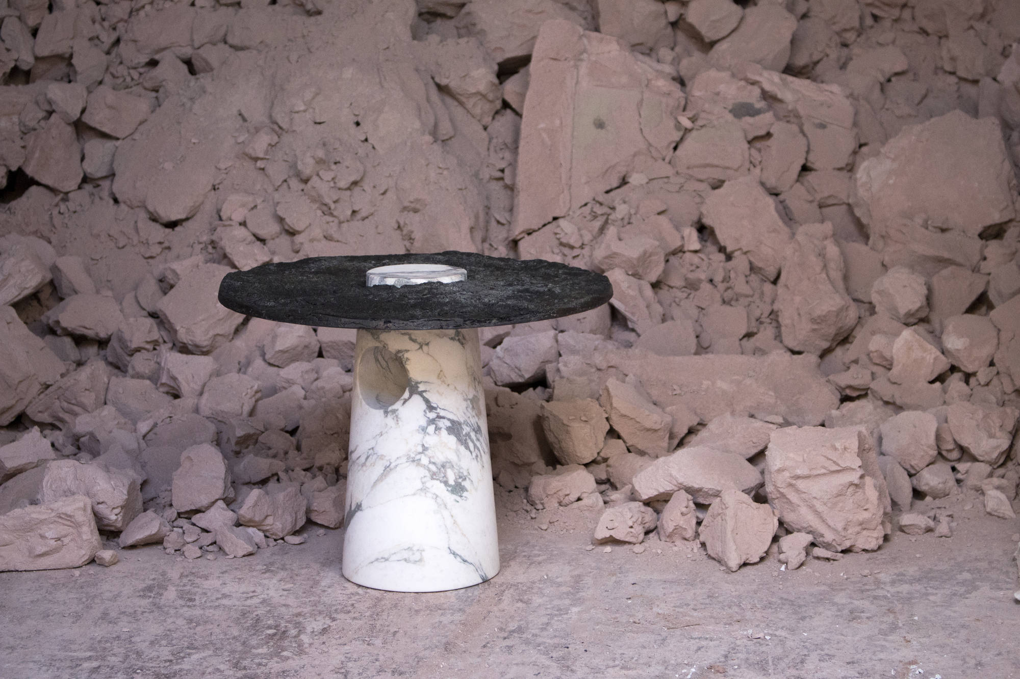 Tavolino in marmo bianco con piano in ardesia nera su sfondo di rocce e detriti.