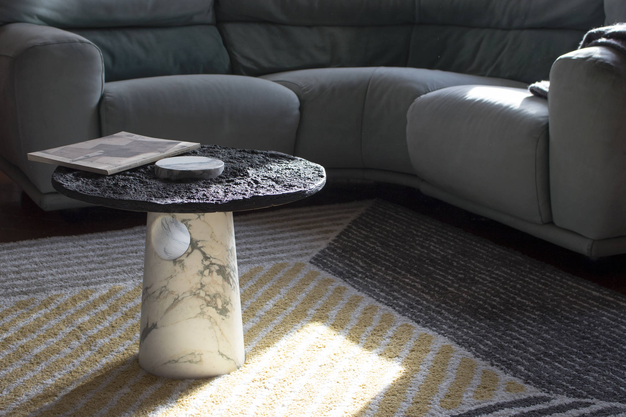 Tavolino moderno in marmo con rivista, accanto a un divano grigio su un tappeto a righe beige e grigie.