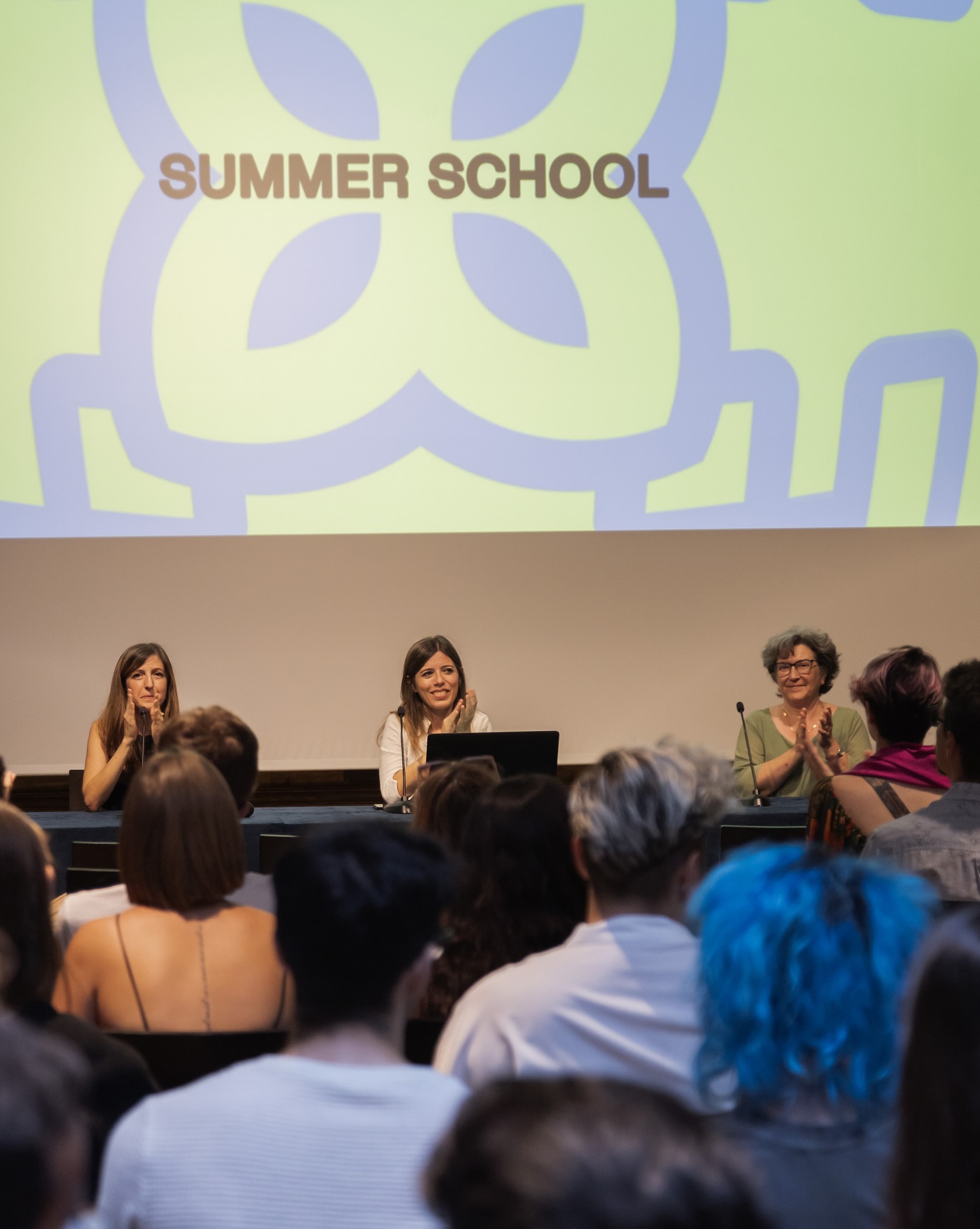 Persone a una conferenza della Summer School, con esperti al tavolo e pubblico attento, schermo con logo sullo sfondo.