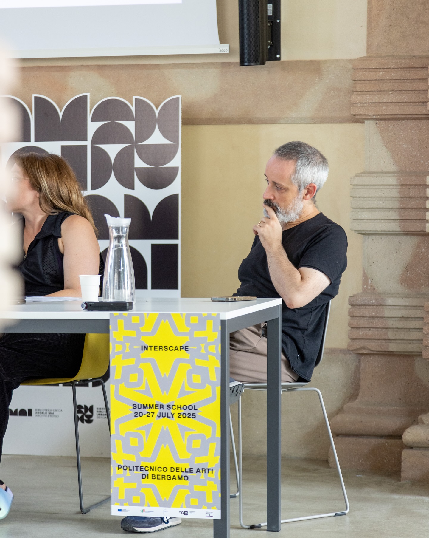Uomo seduto in una conferenza, banner Interscape Summer School 20-27 July 2025, Politecnico delle Arti di Bergamo.