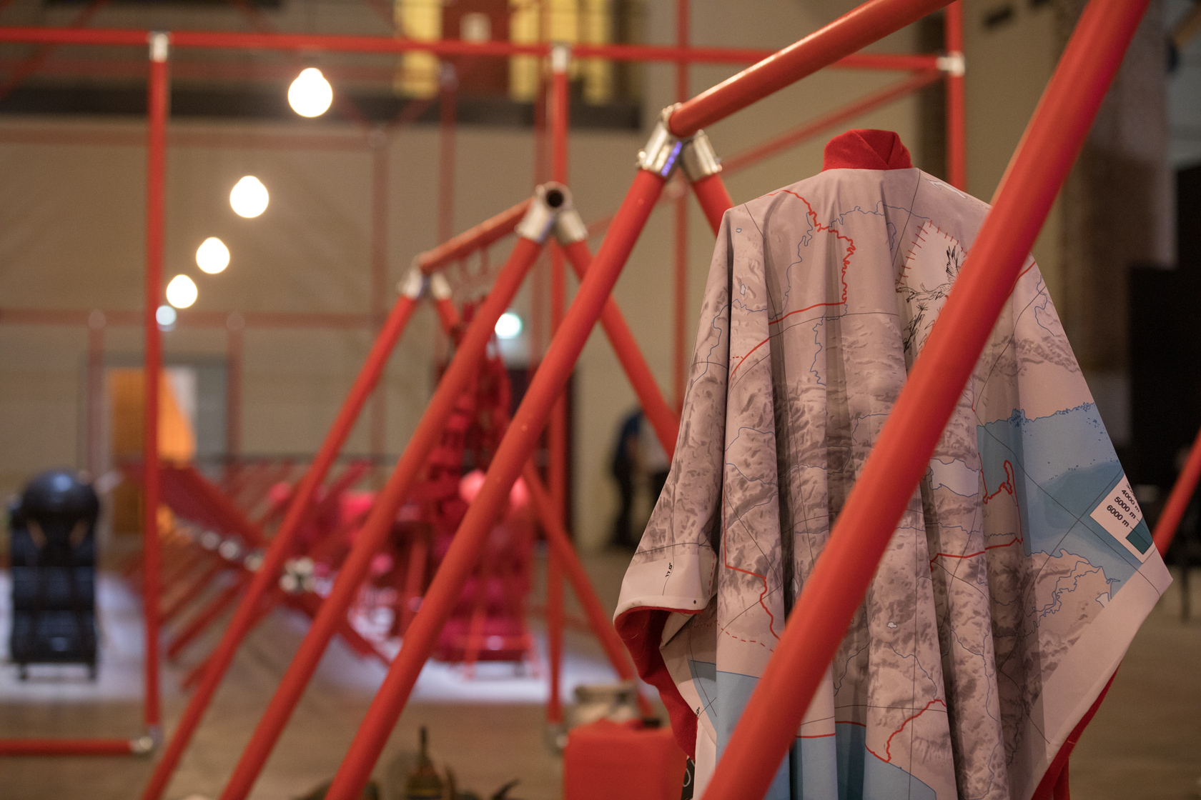 Struttura rossa in tubi di metallo, mappa geografica appesa, illuminazione soffusa in uno spazio espositivo moderno.