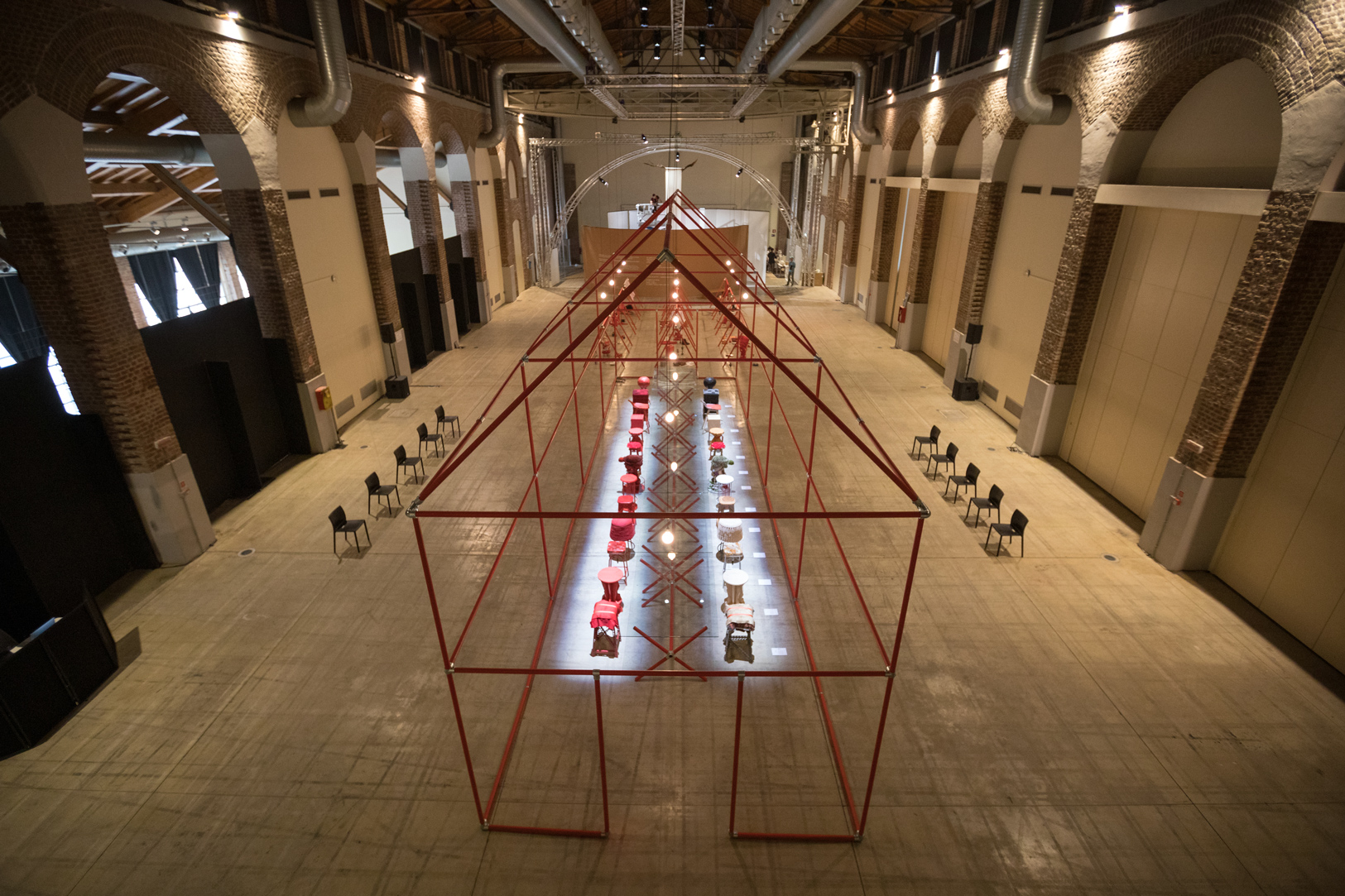 Struttura geometrica rossa con tavoli e sedie colorate all'interno di un ampio spazio industriale con archi in pietra.