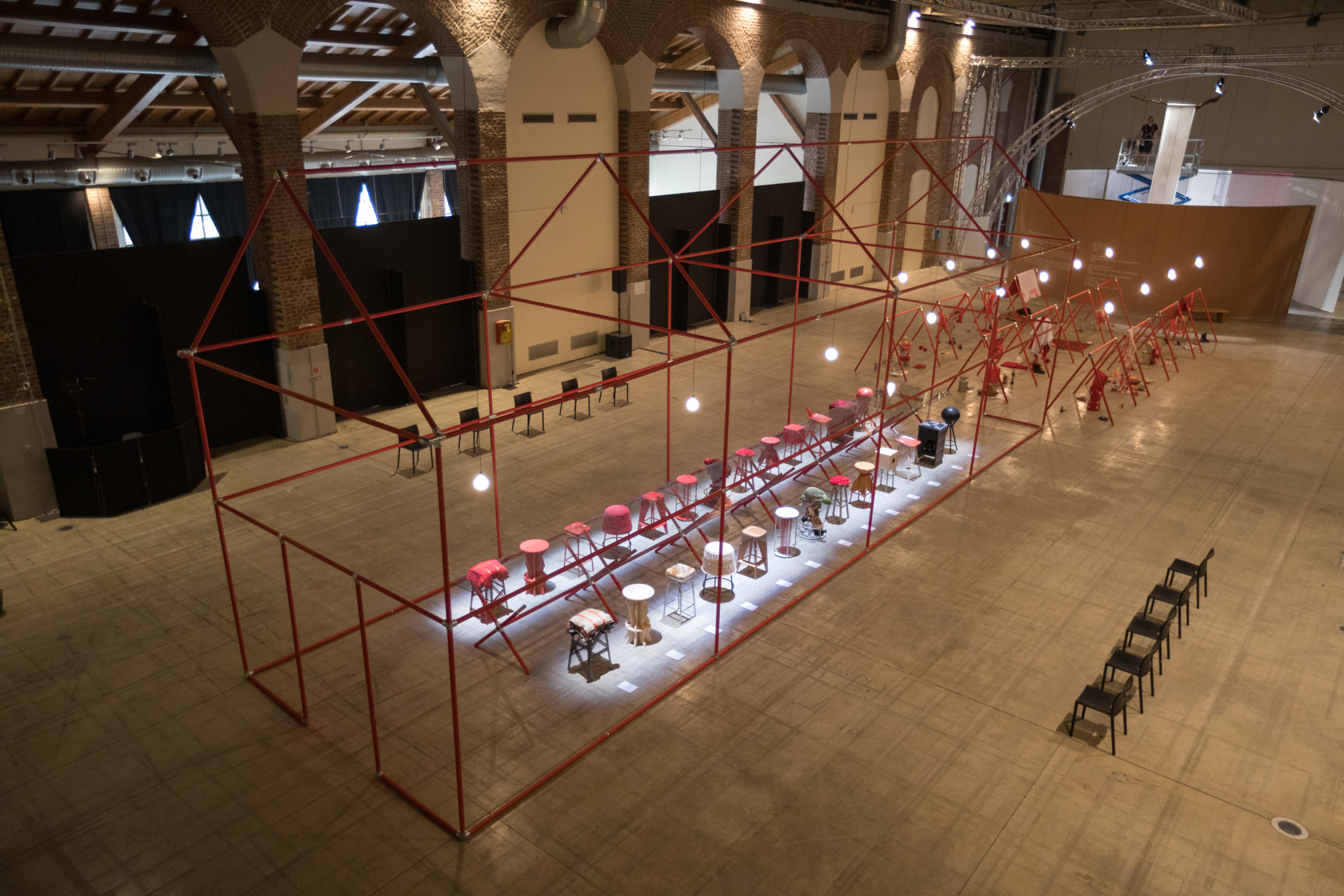 Installazione artistica interna con struttura metallica rossa, sedie colorate illuminate e lampade sospese in uno spazio espositivo.