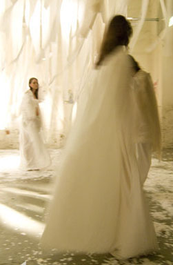 Due persone in abiti bianchi eterei si muovono in una stanza luminosa decorata con nastri bianchi appesi.