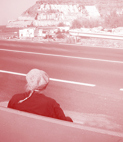 Persona seduta, guarda strada e costa montuosa sullo sfondo, con edifici al sole.