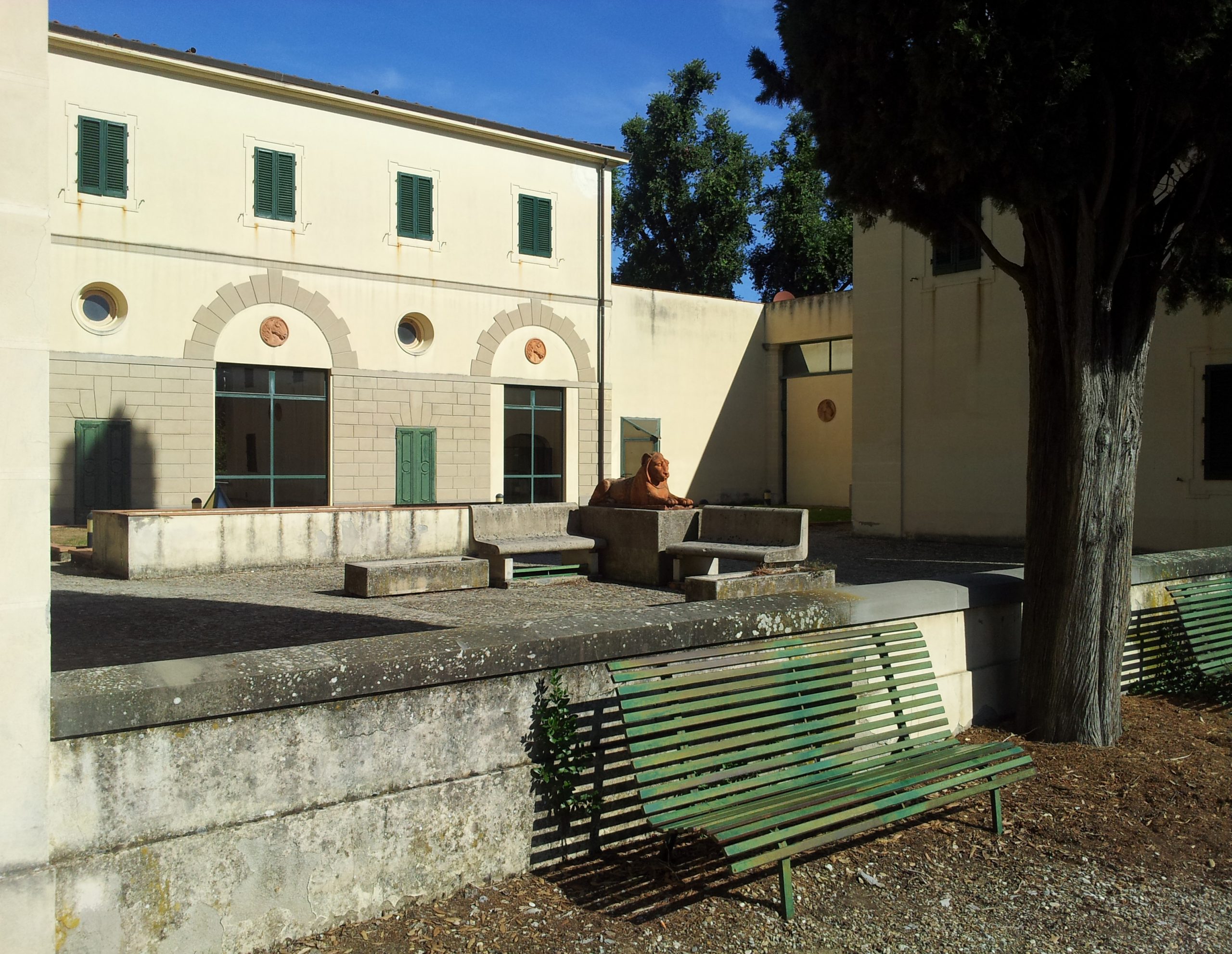 Giardino con panchina verde, statua di leone e edificio storico con finestre verdi e simboli decorativi sulla facciata.