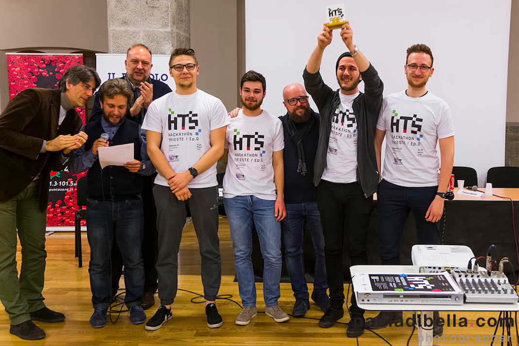Gruppo di partecipanti felici all'Hackathon Trieste, con uno di loro che alza un trofeo mentre altri sorridono.