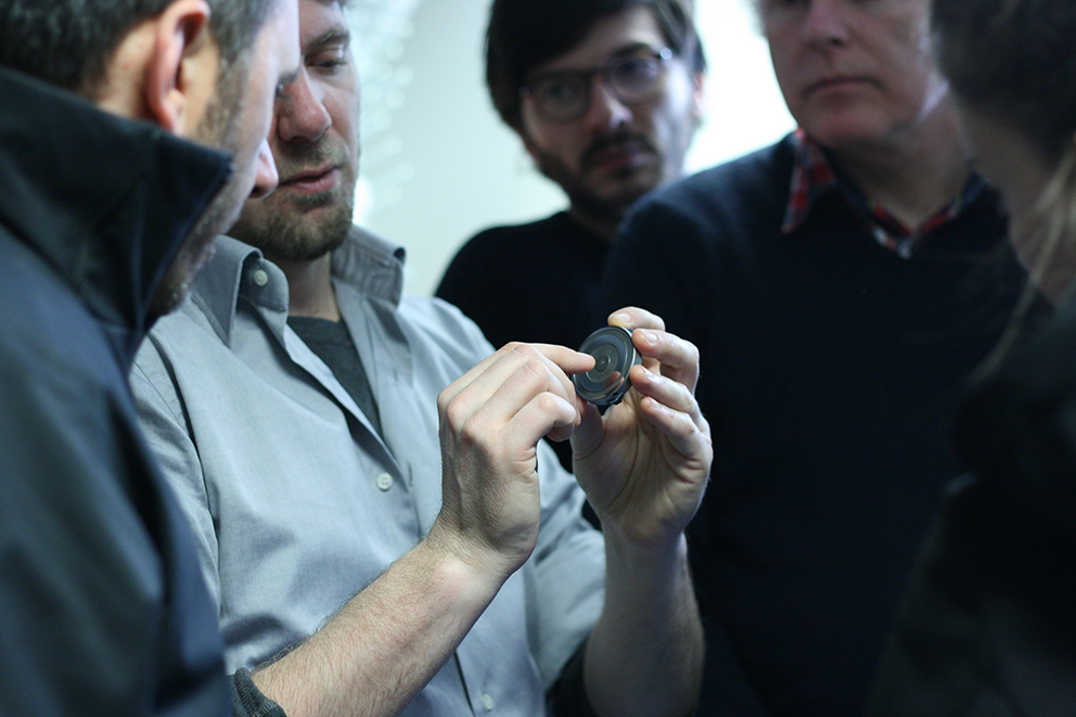 Gruppo di uomini osserva attentamente un piccolo dispositivo tenuto da uno di loro durante una discussione tecnica.