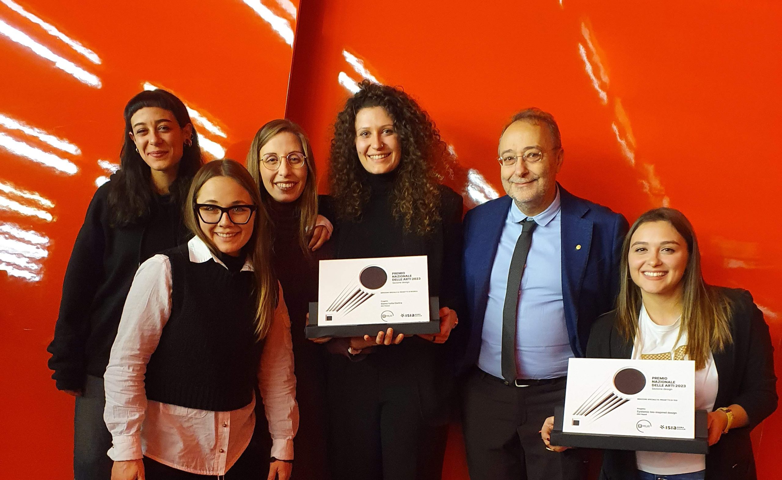 Gruppo di sei persone sorride con premi in mano davanti a una parete arancione brillante.