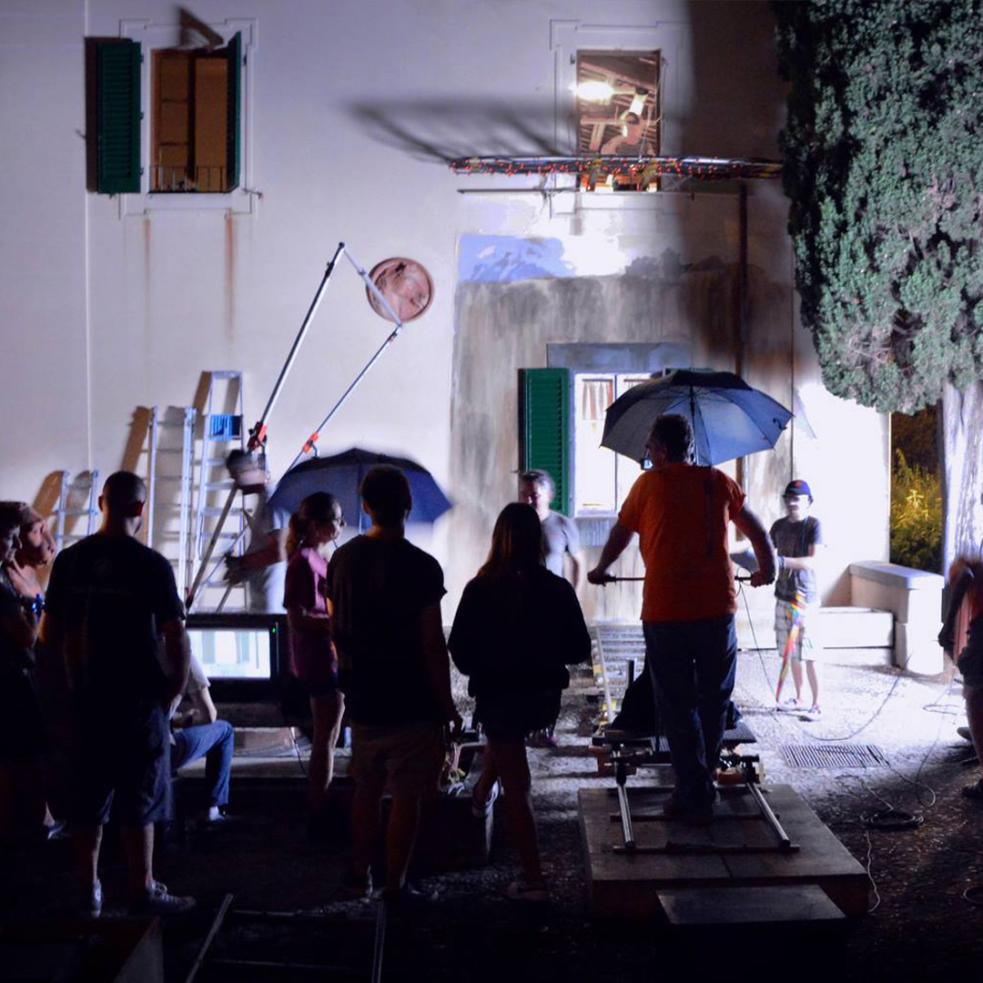 Set cinematografico notturno con tecnici e attrezzature davanti a una casa illuminata, preparazioni per una scena.